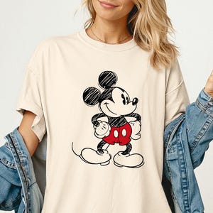 May include: Ivory-colored t-shirt featuring a black and red Mickey Mouse graphic. The cartoon character is drawn in a classic style, with black ears, a white face, and red shorts. The shirt is part of the "Comfort Colors" collection.
