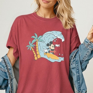 May include: Crimson red t-shirt featuring a graphic of Mickey Mouse surfing a wave. The design includes a palm tree and a yellow surfboard. The shirt is a Comfort Colors brand.