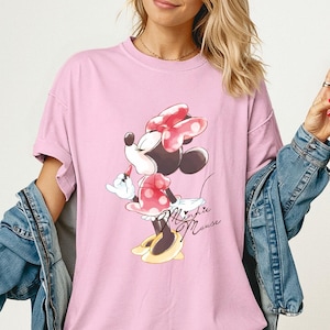 K&ouml;nnte beinhalten: Hellrosa T-Shirt mit Minnie Mouse-Grafik. Das Design zeigt Minnie Mouse in einem roten gepunkteten Kleid und Schleife. Die Worte "Minnie Mouse" sind kursiv geschrieben. Teil der Comfort Colors Blossom-Kollektion.