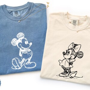 May include: Two Comfort Colors t-shirts, one blue jean and one ivory, each with a sketched cartoon character. The blue shirt features Mickey Mouse, and the ivory shirt features Minnie Mouse. Both shirts have a small gold necklace.