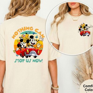 Pode incluir: Camiseta cor marfim com um gr&aacute;fico colorido de Mickey e Minnie Mouse em um carro vermelho, com o texto "NOTHING CAN STOP US NOW!". O design inclui um fundo de p&ocirc;r do sol amarelo, laranja e azul. Uma vers&atilde;o menor do gr&aacute;fico est&aacute; na frente.