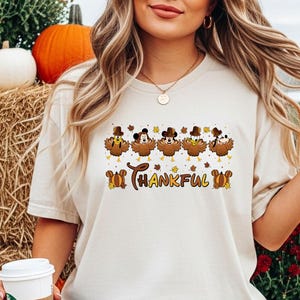 May include: Beige t-shirt with a Thanksgiving-themed graphic. The graphic features cartoon turkeys wearing pilgrim hats and the text "Thankful". The shirt is paired with blue jeans. Pumpkins and hay bales are in the background.