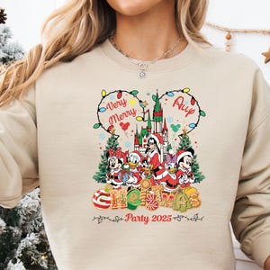May include: Beige crewneck sweatshirt with a colorful Christmas graphic featuring Disney characters. The design includes the text "Very Merry" and "Christmas Party 2025". The graphic showcases a castle, Christmas trees, and candy canes.