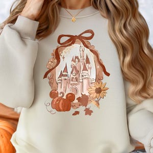 May include: Cream-coloured sweatshirt with a castle design, pumpkins, a sunflower, and autumn leaves. The design includes a brown ribbon and a Mickey Mouse pumpkin. The sweatshirt has long sleeves.
