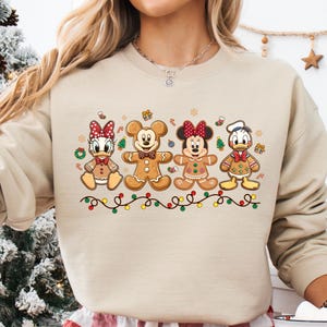 May include: Beige sweatshirt featuring a festive design of Disney characters as gingerbread cookies. The cookies are arranged in a row, with colorful lights below. The sweatshirt is paired with red and white plaid pajama pants.