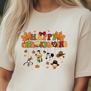 Op de afbeelding: Beige T-shirt met de tekst "HAPPY THANKSGIVING" in kleurrijke letters en herfstbladeren. Onder de tekst staan gezichten van stripfiguren met hoeden en strikken. Het shirt heeft een relaxte pasvorm.