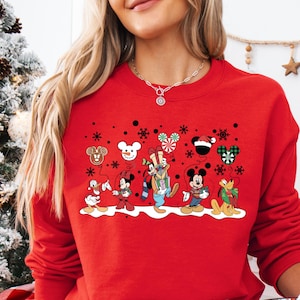 May include: Red Christmas sweatshirt featuring a lineup of Disney characters including Mickey Mouse, Minnie Mouse, Goofy, Pluto, Donald Duck, and Daisy Duck. The characters are depicted in festive attire against a snowy background with snowflakes and other holiday elements. The sweatshirt is a crewneck style with long sleeves.