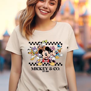 May include: A beige t-shirt featuring a colorful graphic of Mickey Mouse and friends, including Donald Duck, Goofy, and Minnie Mouse. The design is set against a black and white checkered pattern, with the text "MICKEY & CO. EST 1928" below.