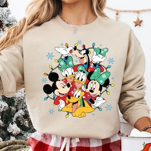 K&ouml;nnte beinhalten: Beiges Sweatshirt mit festlichem Disney-Design. Es zeigt Micky Maus, Minnie Maus, Donald Duck, Goofy und Pluto, umgeben von Weihnachtsbeleuchtung und Schneeflocken. Die Charaktere tragen festliche Kleidung.