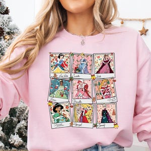 Puede incluir: Una sudadera rosa claro con una cuadr&iacute;cula de nueve retratos de princesas al estilo polaroid. Cada retrato est&aacute; enmarcado con decoraciones festivas navide&ntilde;as. La sudadera es de color rosa claro liso.