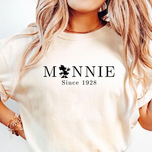 May include: Cream-colored t-shirt with the word "Minnie" in black, featuring a silhouette of a character and the text "Since 1928." The shirt is paired with blue denim shorts. The overall style is casual and comfortable.