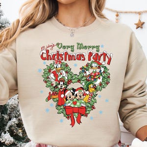 Op de afbeelding: Beige sweatshirt met een kerstthema. De afbeelding toont Mickey Mouse en vrienden, met de tekst "Mickey's Very Merry Christmas Party". Het ontwerp is hartvormig, met sneeuwvlokken en een rode strik.
