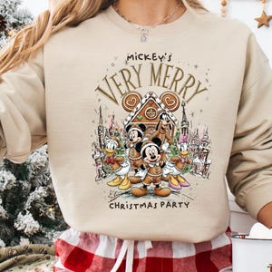 Puede incluir: Sudadera beige con un dise&ntilde;o festivo de Mickey Mouse y sus amigos. El dise&ntilde;o incluye una casa de jengibre y el texto "Mickey's Very Merry Christmas Party". La sudadera se combina con pantalones de pijama a cuadros rojos y blancos.