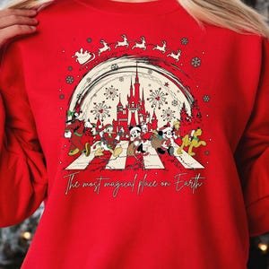 May include: Red Christmas sweatshirt with a white graphic of Mickey Mouse and friends walking across a crosswalk. The graphic includes the words 'The most magical place on Earth' and a silhouette of Cinderella's castle with fireworks. The sweatshirt has a crew neck and long sleeves.