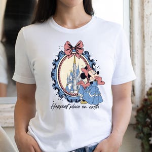 May include: White t-shirt with a graphic of Minnie Mouse standing in front of Cinderella's Castle in a blue and white frame with a pink bow. The text "Happiest place on earth" is printed below the graphic.