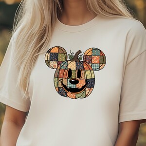 May include: Beige t-shirt featuring a patchwork pumpkin design with Mickey Mouse ears. The pumpkin is composed of various colorful squares and has a smiling face. The shirt is a casual, short-sleeved style.