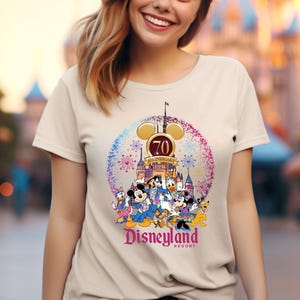 May include: A beige t-shirt with a colorful graphic of Mickey Mouse and friends celebrating Disneyland's 70th anniversary. The graphic features the Disneyland castle, fireworks, and the text "Disneyland Resort".