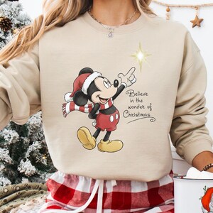 May include: Beige sweatshirt featuring Mickey Mouse in a Santa hat and scarf, pointing at a star. The text "Believe in the wonder of Christmas" is printed on the front. The person is wearing red and white plaid pajama pants.