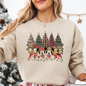 May include: Beige crewneck sweatshirt featuring a festive design with Disney characters, including Mickey and Minnie Mouse, Donald Duck, and Goofy, wearing scarves and hats. Christmas trees and string lights complete the holiday theme.