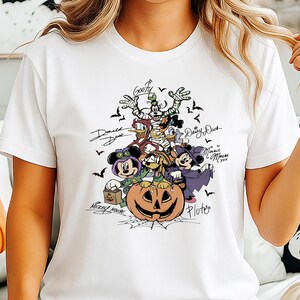 May include: White t-shirt featuring a Halloween-themed graphic of Disney characters, including Mickey Mouse, Donald Duck, and Goofy, surrounding a carved pumpkin. The design includes bats and character signatures.