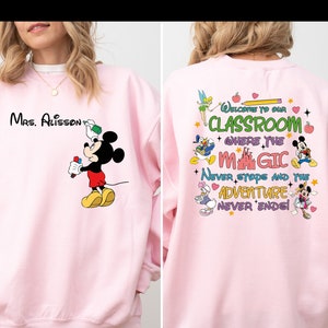 May include: Pink sweatshirt with a Mickey Mouse design on the front and a Disney-themed design on the back. The back design includes the text 'Welcome to our Classroom where the magic never stops and the adventure never ends!'.