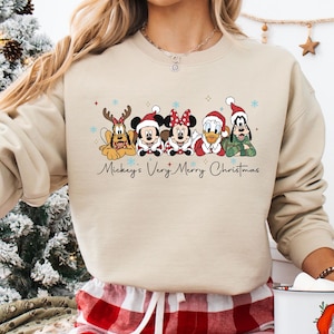 Puede incluir: Sudadera beige con un dise&ntilde;o navide&ntilde;o de personajes de Disney. El texto "Mickey's Very Merry Christmas" est&aacute; escrito debajo. La persona lleva un pantal&oacute;n de pijama a cuadros rojos y blancos y sostiene una taza.