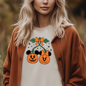 May include: A cream-colored t-shirt featuring two orange pumpkin faces with Mickey Mouse ears. The pumpkins are connected by green vines and a bow. The shirt is worn with a brown cardigan.
