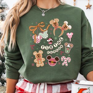 May include: Green sweatshirt with a festive design featuring gingerbread men, Mickey Mouse ears, candy canes, and the phrase "Tis the Season." The design includes pink, white, and brown accents. A white mug with a Santa Claus graphic is held in the hand.