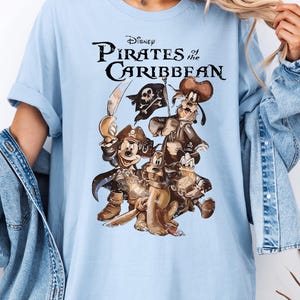 Pode incluir: Camiseta azul claro com uma estampa gr&aacute;fica de Mickey Mouse, Pateta e Donald Duck vestidos de piratas. O texto "Disney Pirates of the Caribbean" est&aacute; impresso acima dos personagens.