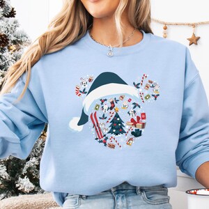 May include: A light blue sweatshirt featuring a festive design. The graphic includes a dark blue Santa hat, a Christmas tree, skis, and wrapped gifts. The design is surrounded by candy canes and other holiday-themed elements.