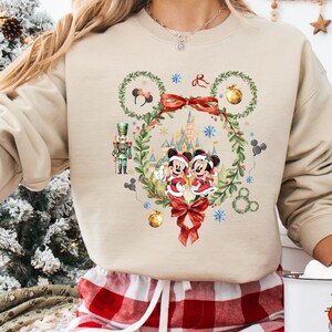 May include: Beige sweatshirt featuring a festive Mickey and Minnie Mouse design. The graphic includes a wreath, castle, and snowflakes. The sweatshirt is paired with red and white plaid pajama pants. A white mug with a Santa Claus graphic is also visible.