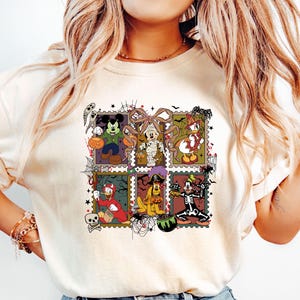 以下が含まれることがあります： クリーム色のTシャツ。ハロウィーンをテーマにしたカラフルなグラフィックが特徴です。グラフィックは、ハロウィーンのコスチュームを着た様々な漫画のキャラクターをグリッドパターンで表示しています。デザインには、幽霊、カボチャ、クモの巣が含まれています。