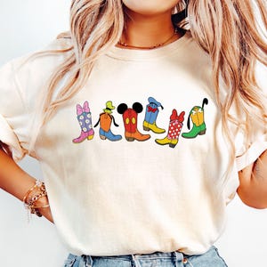 May include: Cream-colored t-shirt featuring a row of cartoon cowboy boots in various colors and designs. The boots include floral patterns, Mickey Mouse ears, and polka dots. The boots are arranged horizontally across the shirt.