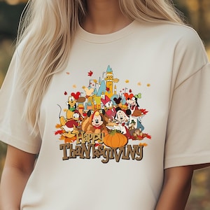 May include: Beige t-shirt featuring a colorful graphic of Disney characters gathered around a pumpkin with the text "Happy Thanksgiving". The design includes a castle and autumn leaves.
