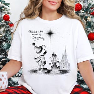 May include: White Christmas-themed t-shirt with a black and white illustration of cartoon characters and the text "Believe in the wonder of Christmas." The design includes a star, a castle, and two characters wearing winter attire.