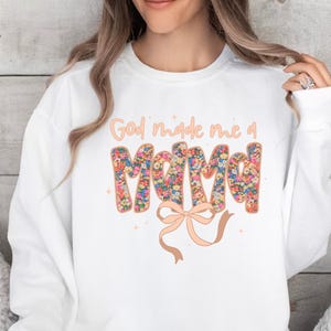 Pu&ograve; includere: Felpa bianca con la scritta "God made me a Mama" in un design floreale. La parola "Mama" &egrave; riempita di fiori colorati. Un fiocco color pesca &egrave; sotto il testo. La felpa &egrave; in stile casual.