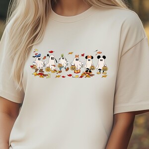 May include: Beige t-shirt featuring a Halloween-themed graphic. The graphic depicts cartoon ghosts dressed as popular characters, each holding a pumpkin-shaped candy bucket. Autumn leaves are scattered around the ghosts.