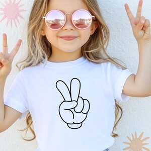 K&ouml;nnte beinhalten: Ein wei&szlig;es T-Shirt mit einem schwarzen Cartoon-Friedenszeichen. Die Person auf dem Bild tr&auml;gt rosa get&ouml;nte runde Sonnenbrillen und macht mit ihren Fingern ein Friedenszeichen. Der Hintergrund ist eine wei&szlig;e Wand mit gelben und rosa sonnenf&ouml;rmigen Designs.