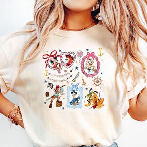 May include: A white t-shirt with a colorful graphic print featuring various Disney characters, including Mickey Mouse, Minnie Mouse, Donald Duck, Goofy, and Pluto. The graphic includes hearts, stars, anchors, and other nautical elements. The text "Wishes Do Come True" is included in the graphic.