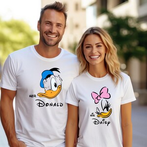 May include: White t-shirts with cartoon graphics of Donald and Daisy Duck. The Donald shirt has a blue hat and the text "Her Donald". The Daisy shirt has a pink bow and the text "His Daisy". Both shirts are paired with blue jeans.