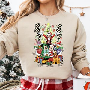 May include: Beige crewneck sweatshirt featuring a festive graphic of Disney characters, including Mickey Mouse, Donald Duck, and Goofy, celebrating Christmas. The design includes a Christmas tree, presents, and candy canes. The sweatshirt has a relaxed fit.