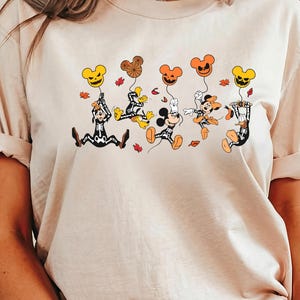 May include: Beige t-shirt with a Halloween graphic. The design shows cartoon characters in costumes, holding pumpkin-shaped balloons. The balloons are in different colours, and there are autumn leaves scattered around the characters.