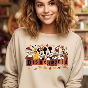 May include: Beige sweatshirt featuring a Disney World graphic with cartoon characters like Mickey and Minnie Mouse, Donald Duck, and Goofy. The graphic includes the text "Disneyworld" in a fall-themed design with leaves.
