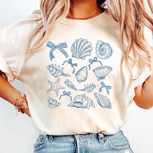Pu&ograve; includere: Una maglietta bianca con un design di conchiglie e stelle marine blu. Il design presenta varie conchiglie, stelle marine e fiocchi.