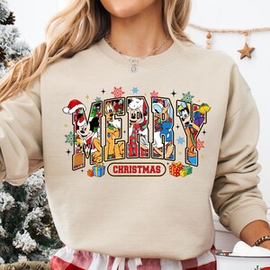 May include: A beige sweatshirt with the word "MERRY CHRISTMAS" in colorful block letters. The letters are filled with cartoon characters, snowflakes, and gift boxes. The sweatshirt is part of a holiday-themed collection.
