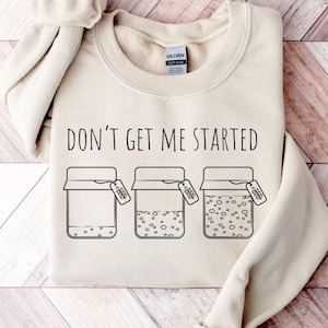 May include: A cream-colored sweatshirt with the phrase "DON'T GET ME STARTED" printed in black. Below the text are three jar illustrations with varying contents and tags. The sweatshirt is folded on a wooden surface.