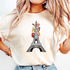 May include: Cream-colored t-shirt featuring a black Eiffel Tower design with colorful cartoon princess figures. The graphic is centered on the front of the shirt. The shirt has short sleeves and a crew neck.