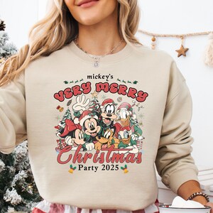 Puede incluir: Sudadera beige con un gr&aacute;fico colorido de Mickey Mouse, Minnie Mouse, Donald, Goofy y Pluto celebrando la Navidad. El texto "mickey's VERY MERRY Christmas Party 2025" est&aacute; impreso encima y debajo de los personajes.