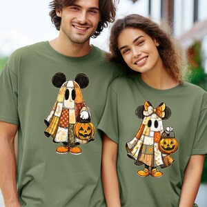 May include: Two olive green t-shirts featuring a cartoon ghost design with Mickey Mouse ears, a patchwork cloak, and a jack-o'-lantern. The shirts are worn by a man and a woman, perfect for Halloween.