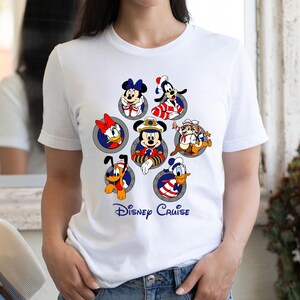 May include: White t-shirt featuring a colorful graphic of Disney characters, including Mickey Mouse, Minnie Mouse, Donald Duck, and Goofy, in circular frames. The words "Disney Cruise" are printed in blue script below the characters. The shirt is paired with denim shorts.